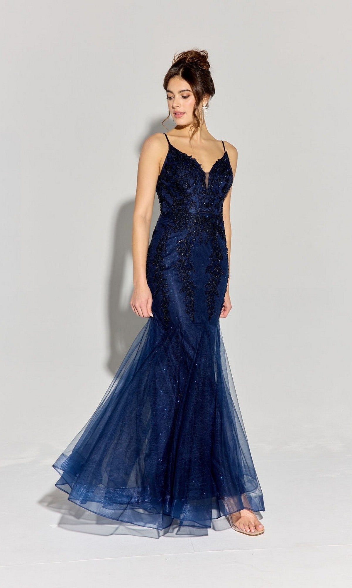 A woman wears the Eureka 9998 long navy prom dress with a mermaid godet silhouette, thin straps, and lace details, standing against a plain light gray background.